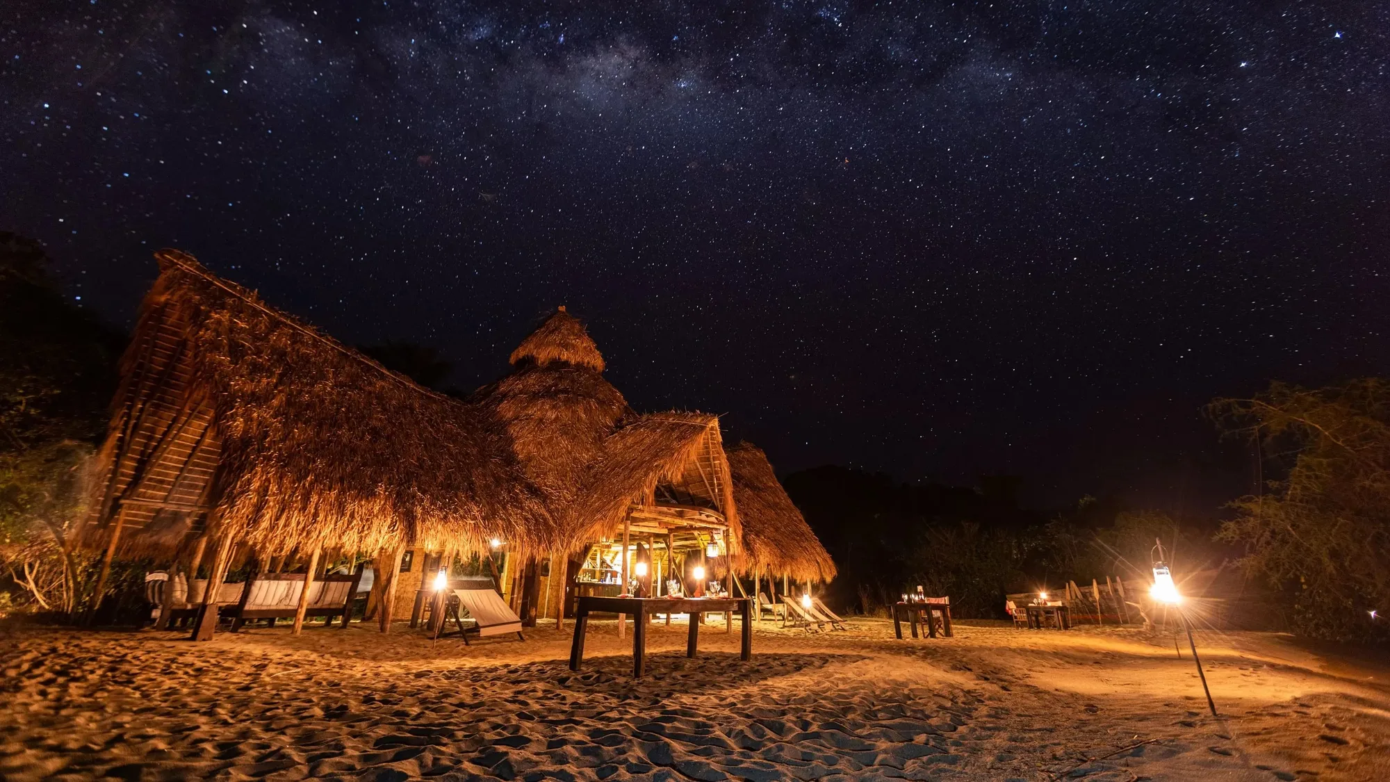 A thatched lodge on a Lake Tanganyika beach lit by lanterns under the Milky Way