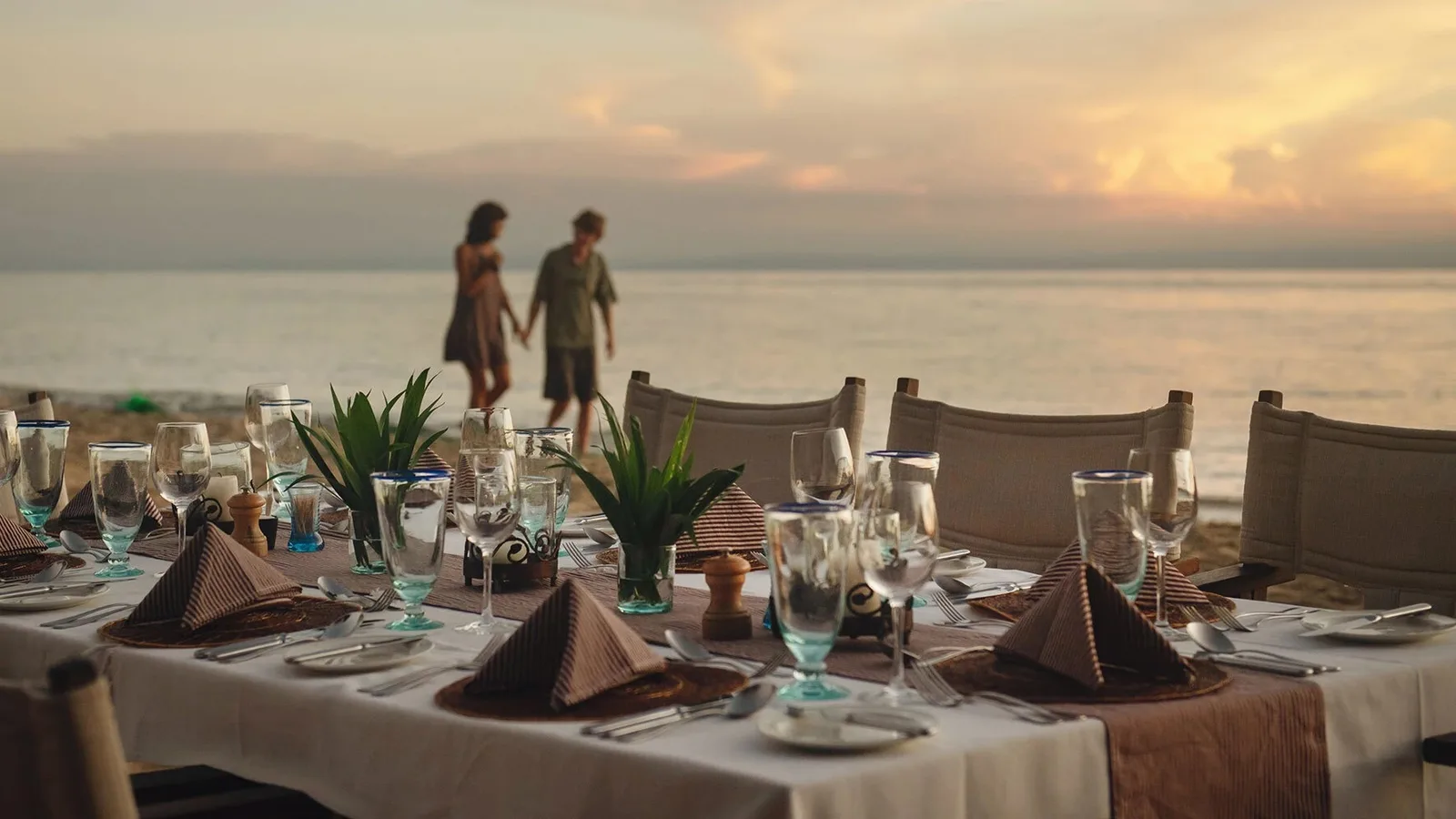 A couple walking on the beach beside a dinner table laid on the shore of Lake Tanganyika at sunset